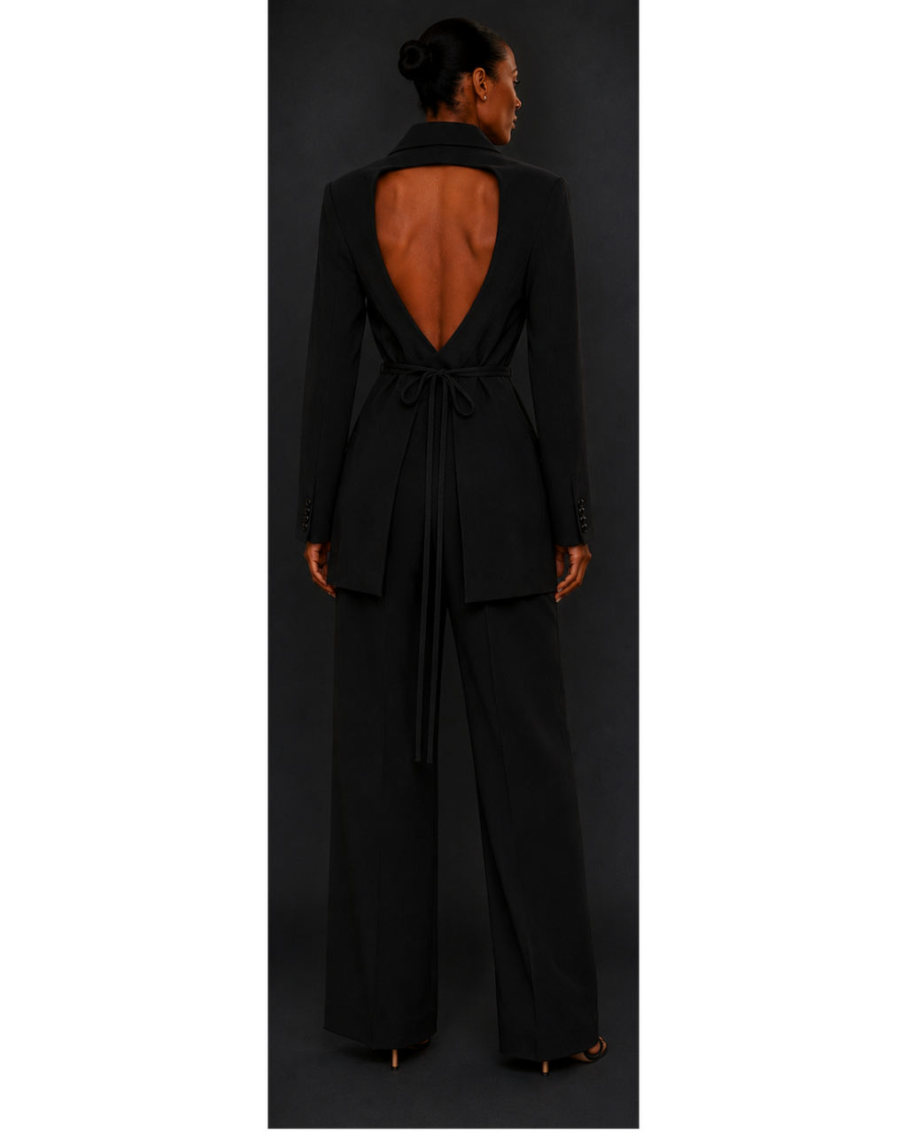 Black open-back blazer and black trousers worn by a model on a dark background