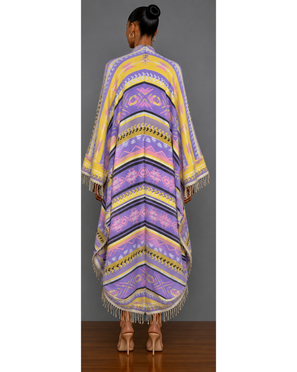 Back view of a woman wearing a colorful patterned blanket wrap coat with fringe against a gray background