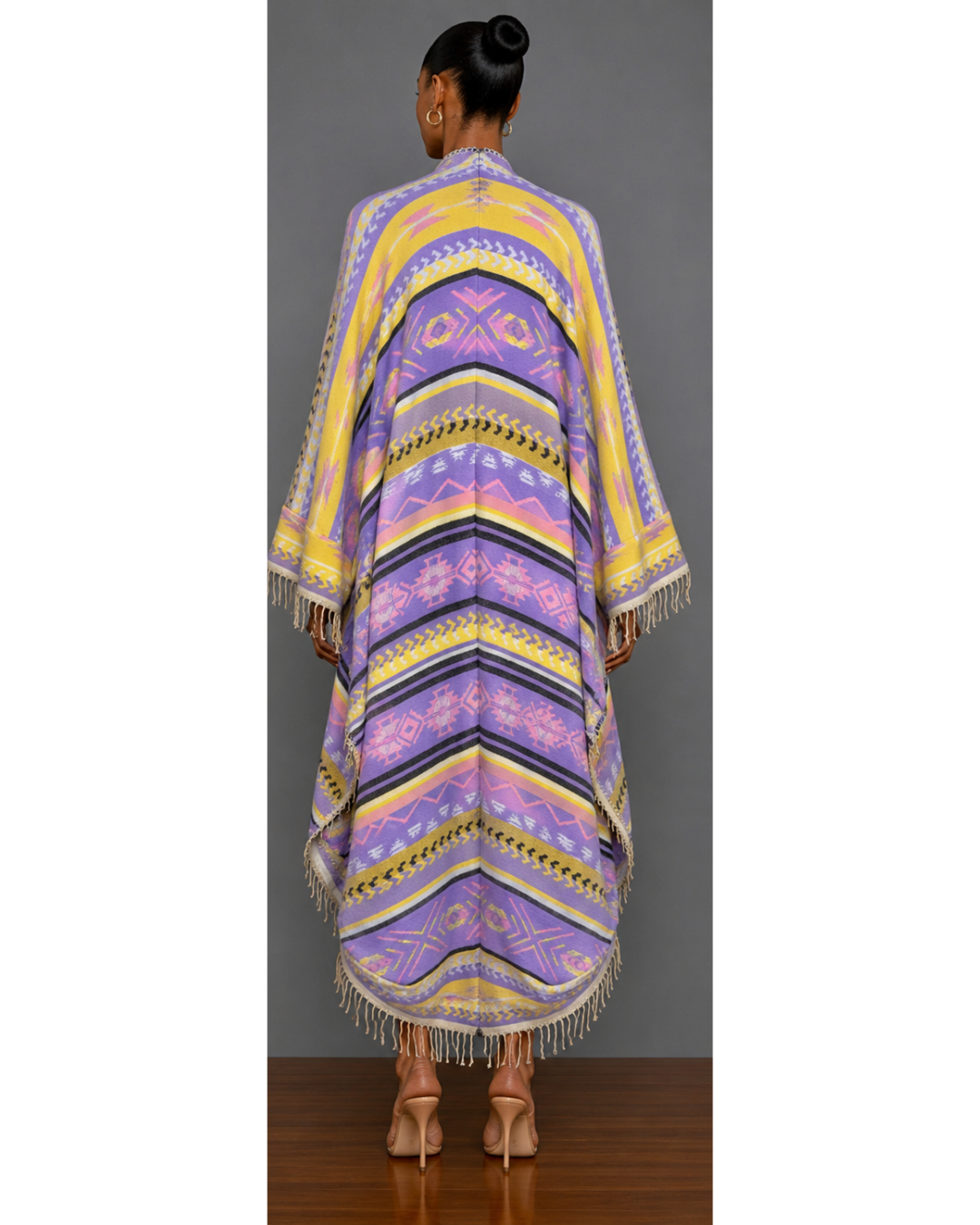 Back view of a woman wearing a colorful patterned blanket wrap coat with fringe against a gray background