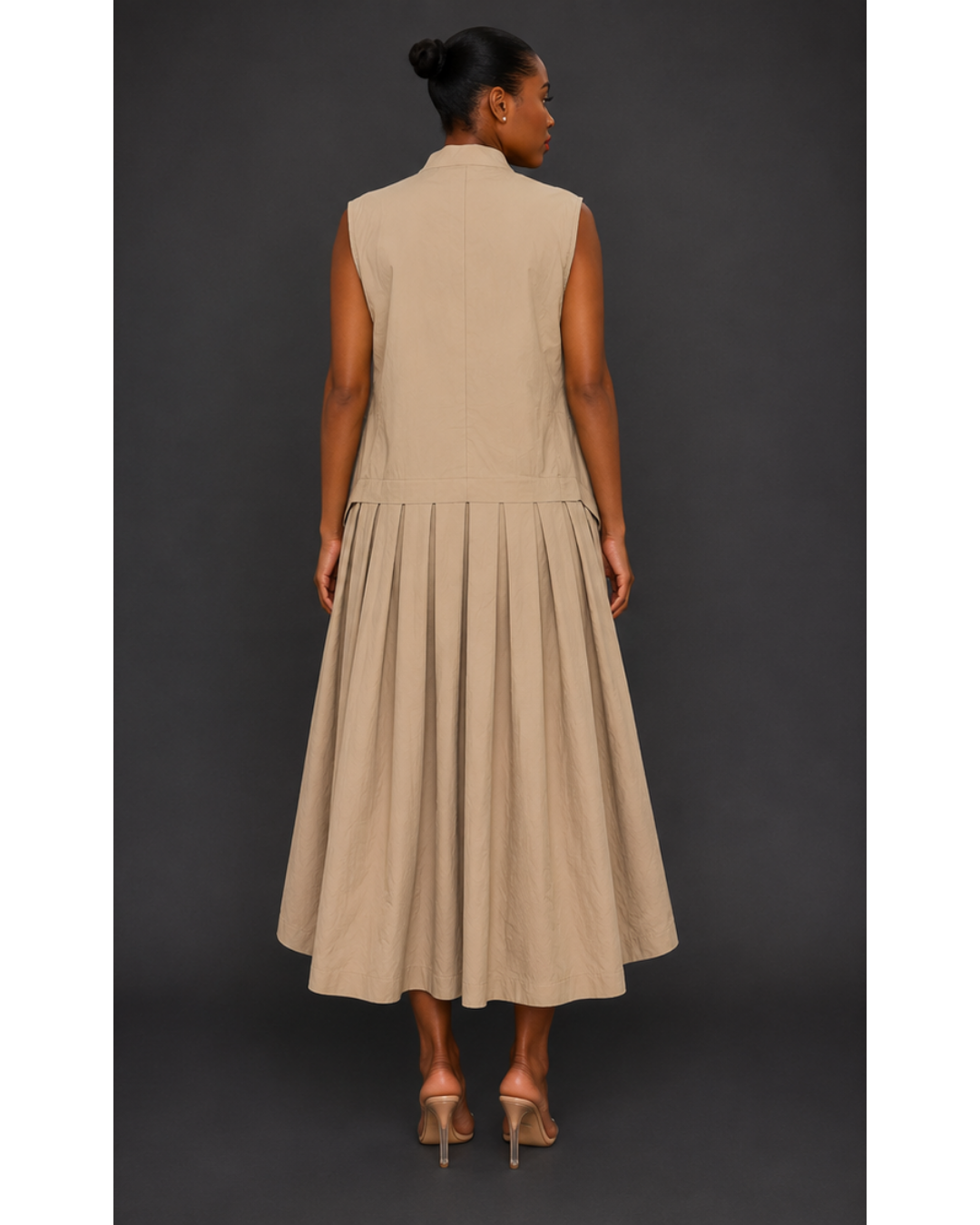 Back view of a woman wearing a beige sleeveless pleated maxi dress against a dark background