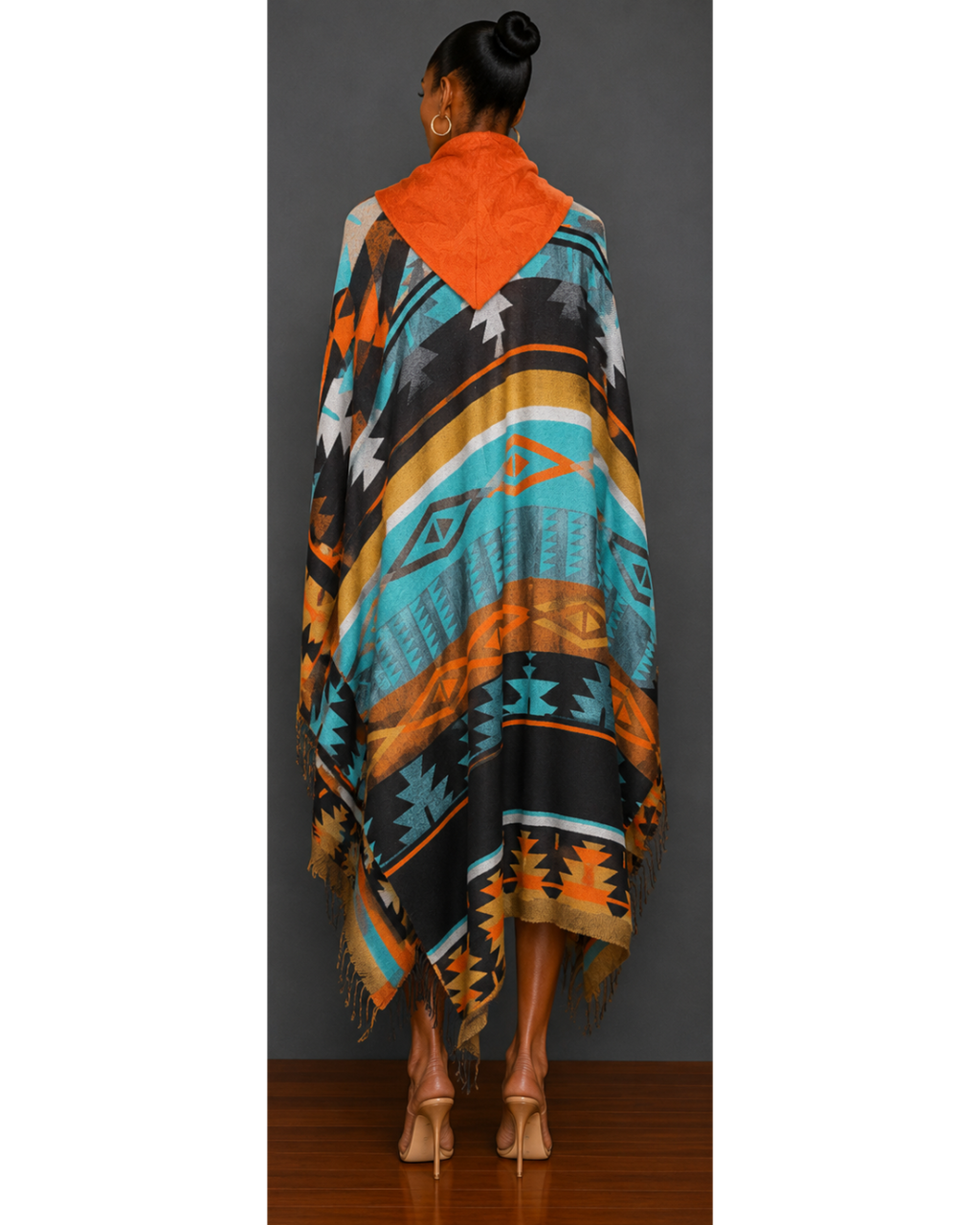 Back view of woman wearing a teal, orange, brown, gold, and black patterned blanket wrap coat with orange hood and fringe hem details