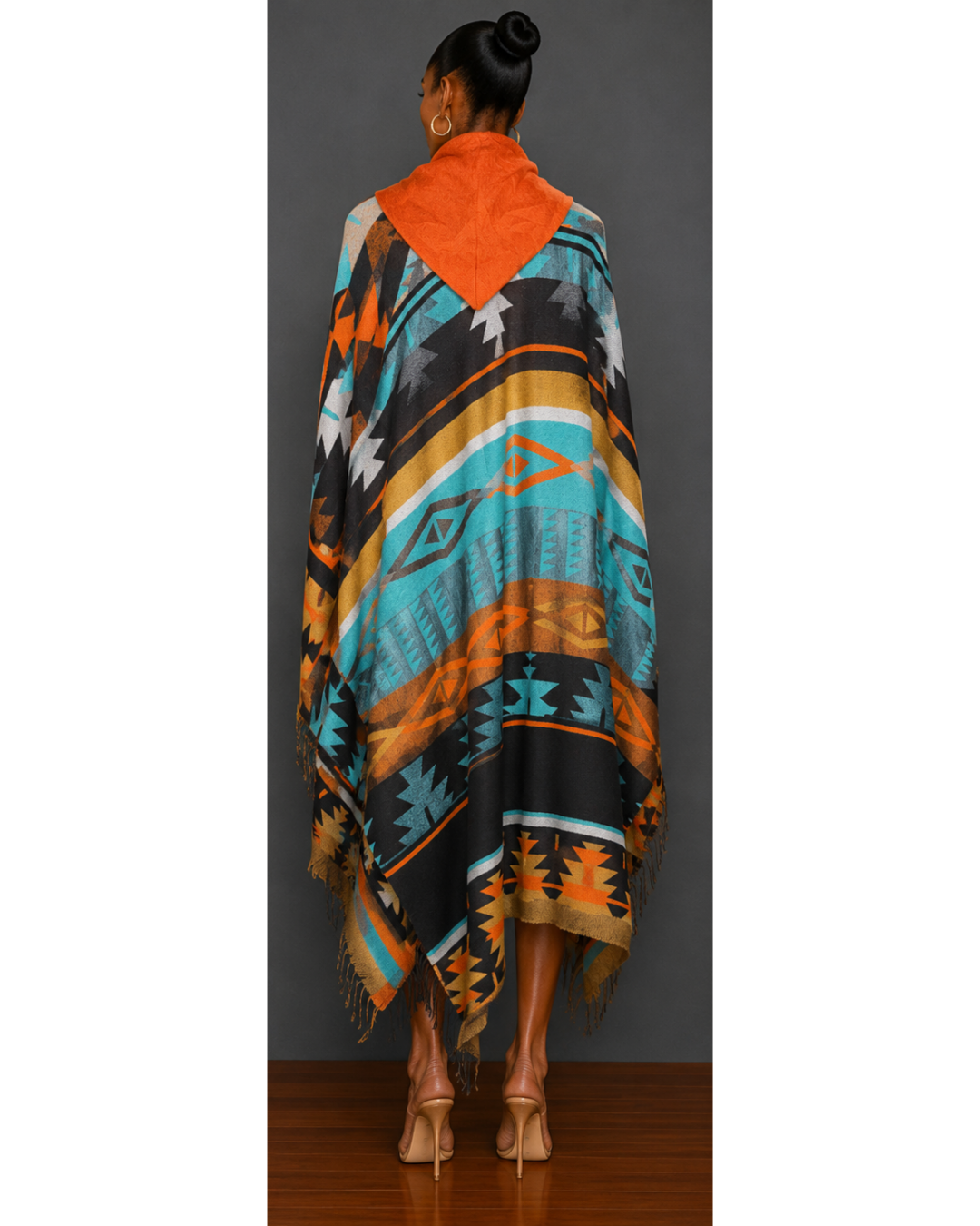 Back view of woman wearing a teal, orange, brown, gold, and black patterned blanket wrap coat with orange hood and fringe hem details