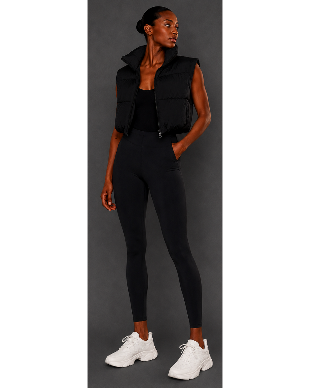 Woman wearing a black sleeveless jacket, black leggings, and white sneakers on a gray background