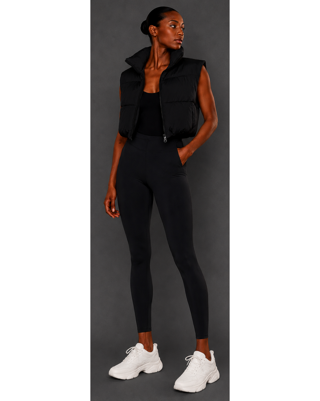Woman wearing a black sleeveless jacket, black leggings, and white sneakers on a gray background